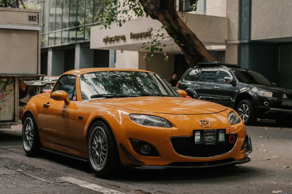 Orange Mazda sports car parked on a city street in front of office buildings, showcasing urban lifestyle.